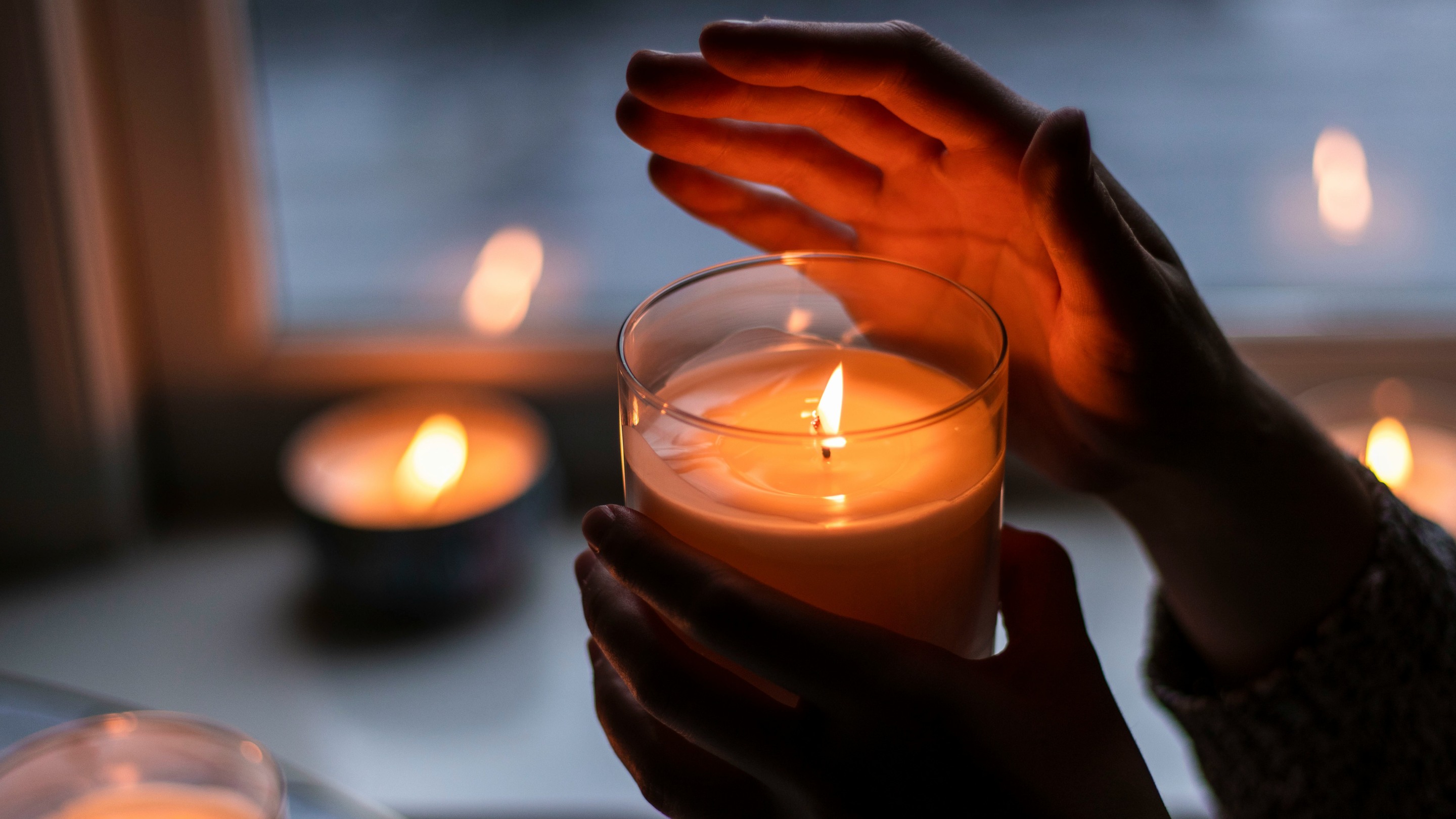 A pair of hands holding a lit candle in a glass jar, with other candles glowing softly in the background near a window.