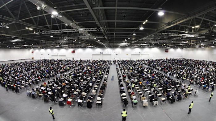 Thousands of people sit at individual desks in rows taking exams in a large hall, with exam invigilators standing next to them