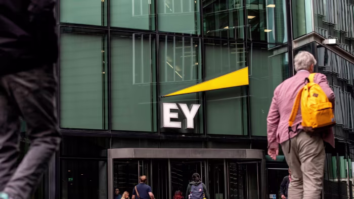 A large "EY" sign with a yellow triangle above the entrance to an office building, people walking past outside.
