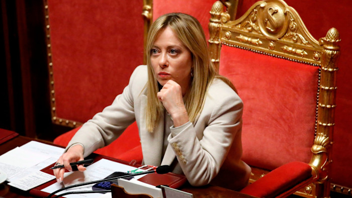Giorgia Meloni seated at a red and gold chair in the Senate, appearing thoughtful with her chin resting on her hand.