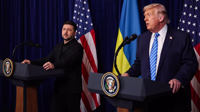 Volodymyr Zelenskyy listens as Donald Trump speaks at podiums with US and Ukrainian flags behind them during a press conference.
