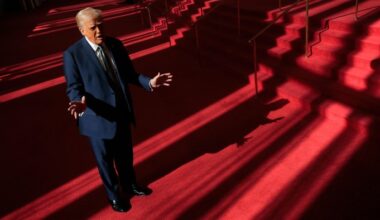 Donald Trump stands in a sunlit foyer with red carpet, gesturing as he speaks to the media near a staircase.