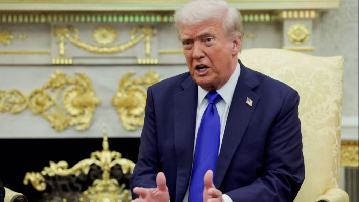 Donald Trump speaks while seated in the Oval Office, gesturing with his hands, with ornate gold decor in the background.