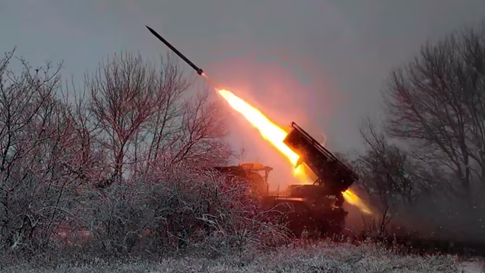 A Russian "Grad" multiple rocket launcher fires a rocket amid trees and winter vegetation, with bright flames visible.