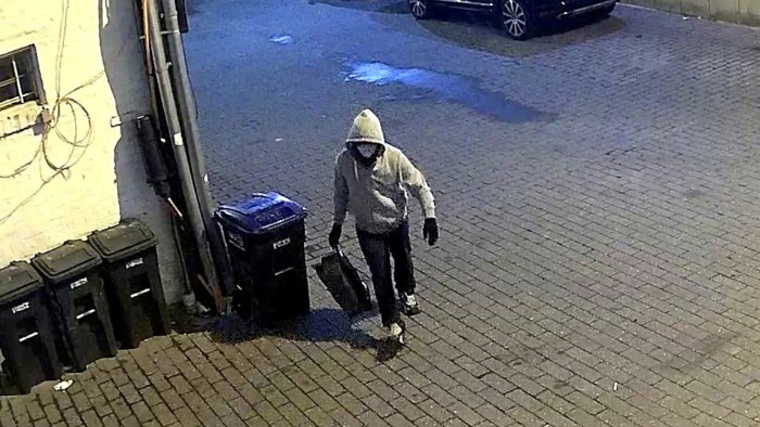 A masked individual in a hooded sweatshirt walks along a brick alley carrying a bag, as captured by a surveillance camera.