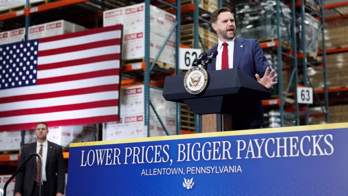 Vice-president JD Vance speaks at a podium in a warehouse with an American flag and a sign reading "Lower Prices, Bigger Paychecks."