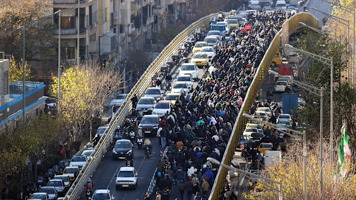 A large crowd of people march along a bridge in Tehran, filling one side while cars are jammed on the other during a protest.