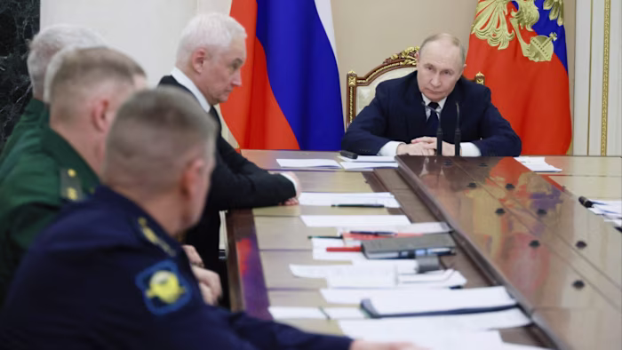 Vladimir Putin sits at the head of a long table during a meeting, with officials and documents in front of him. Russian flags are visible behind him.