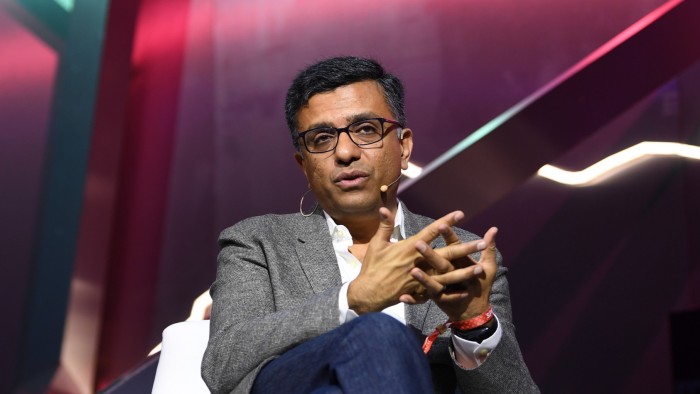 TS Anil speaks on stage, gesturing with his hands, wearing a headset microphone and business attire.