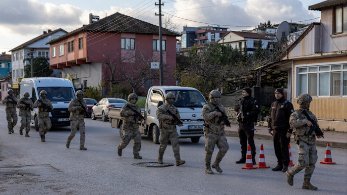 Turkish gendarmerie special forces in camouflage gear and helmets walk past parked vehicles and two uniformed officers near traffic cones on a residential street.