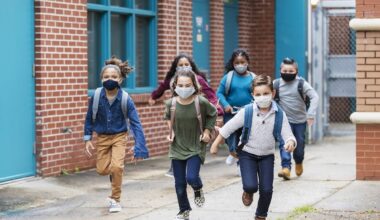 A group of six elementary school students running outside the school building, carrying backpacks, and wearing face masks. They are back to school during the covid-19 pandemic. The boys and girls are 7 to 10 years old.