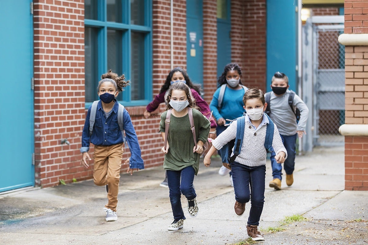 A group of six elementary school students running outside the school building, carrying backpacks, and wearing face masks. They are back to school during the covid-19 pandemic. The boys and girls are 7 to 10 years old.