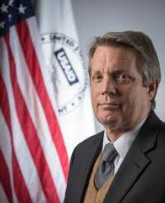 A headshot of a man in a suit looking at the camera, in front of the American flag and the USAID logo.