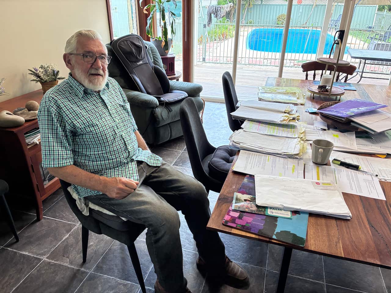 A photo of an elderly man with white hair and a beard, sitting in his home office surrounded by notebooks and documents.