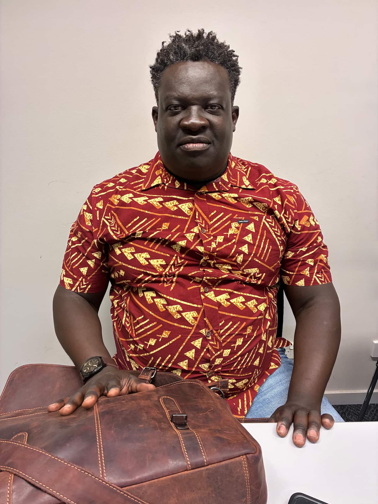 A middel-aged black man wearing a red and yellow patterned shirt sits in an office.