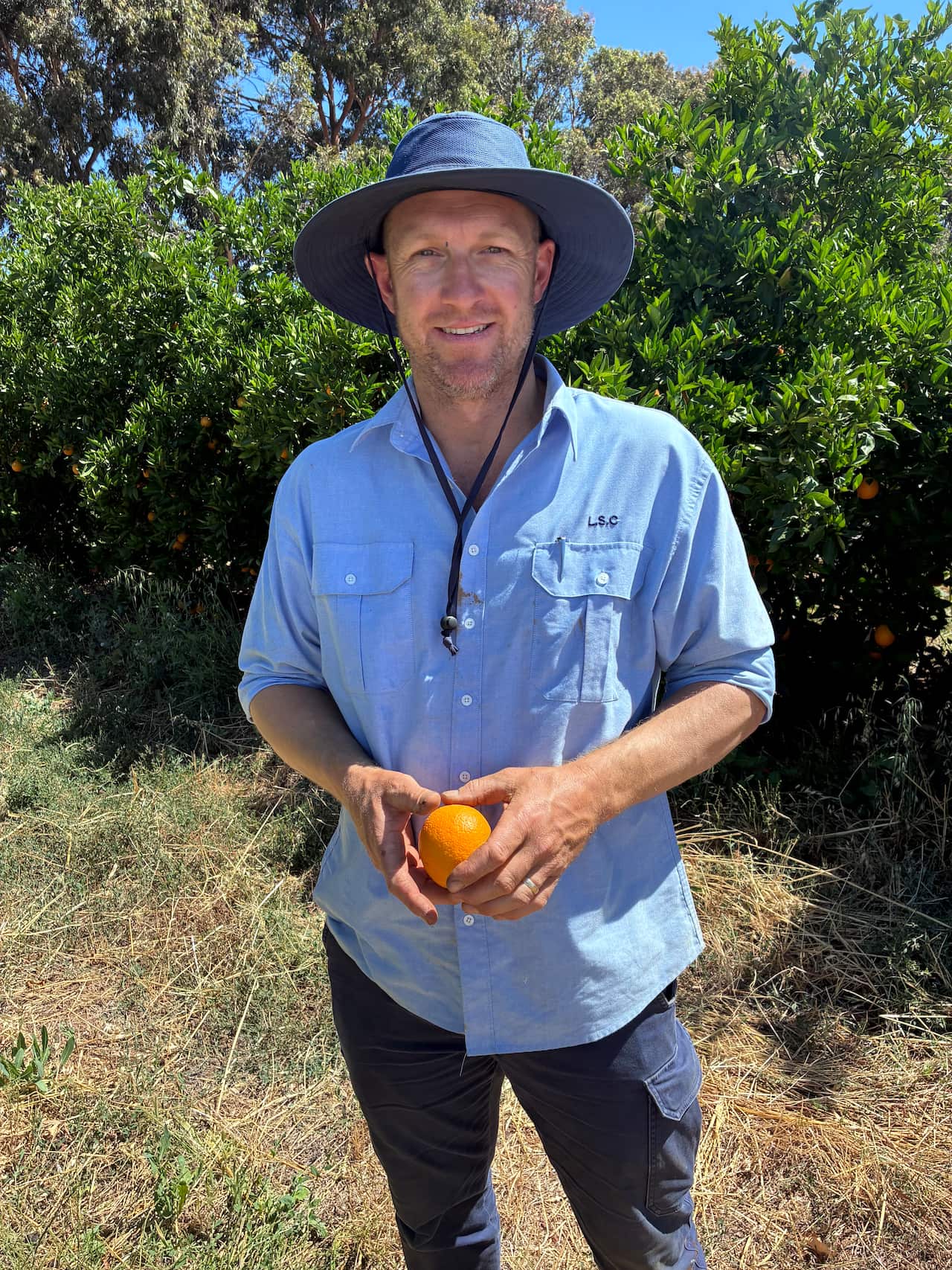 A middle-aged man stands outside among trees and dry grass, holding an orange.