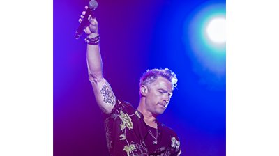 A man stands on stage in a black t-shirt with a floral print, holding his arm aloft with a microphone in hand