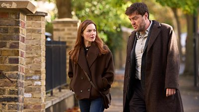 A man and a woman (Strike and Robin) walk together outdoors on a tree-lined road, both in brown coats