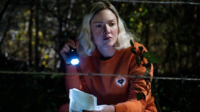 A woman (Robin) in a blonde wig and orange tracksuit crouches down by some wire at night, holding a torch and a piece of paper