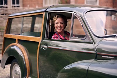 A woman (Claudie Blakley playing Mrs Sullivan) sits in a vintage car smiling 