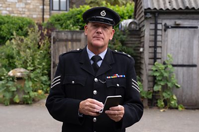 A man in a police sergeant uniform (John Burton as Sergeant Goodfellow in Father Brown) looks on with a notepad 
