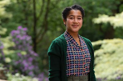 A woman in a colourful shirt and a cardigan (Ruby May-Martinwood as Brenda in Father Brown) looks on smiling in a garden-setting 