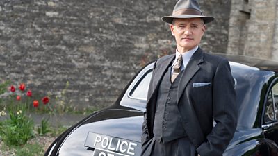 Tom Chambers in character as Inspector Sullivan. He stands beside an old-fashioned police car, wearing a three-piece suit and a hat. 