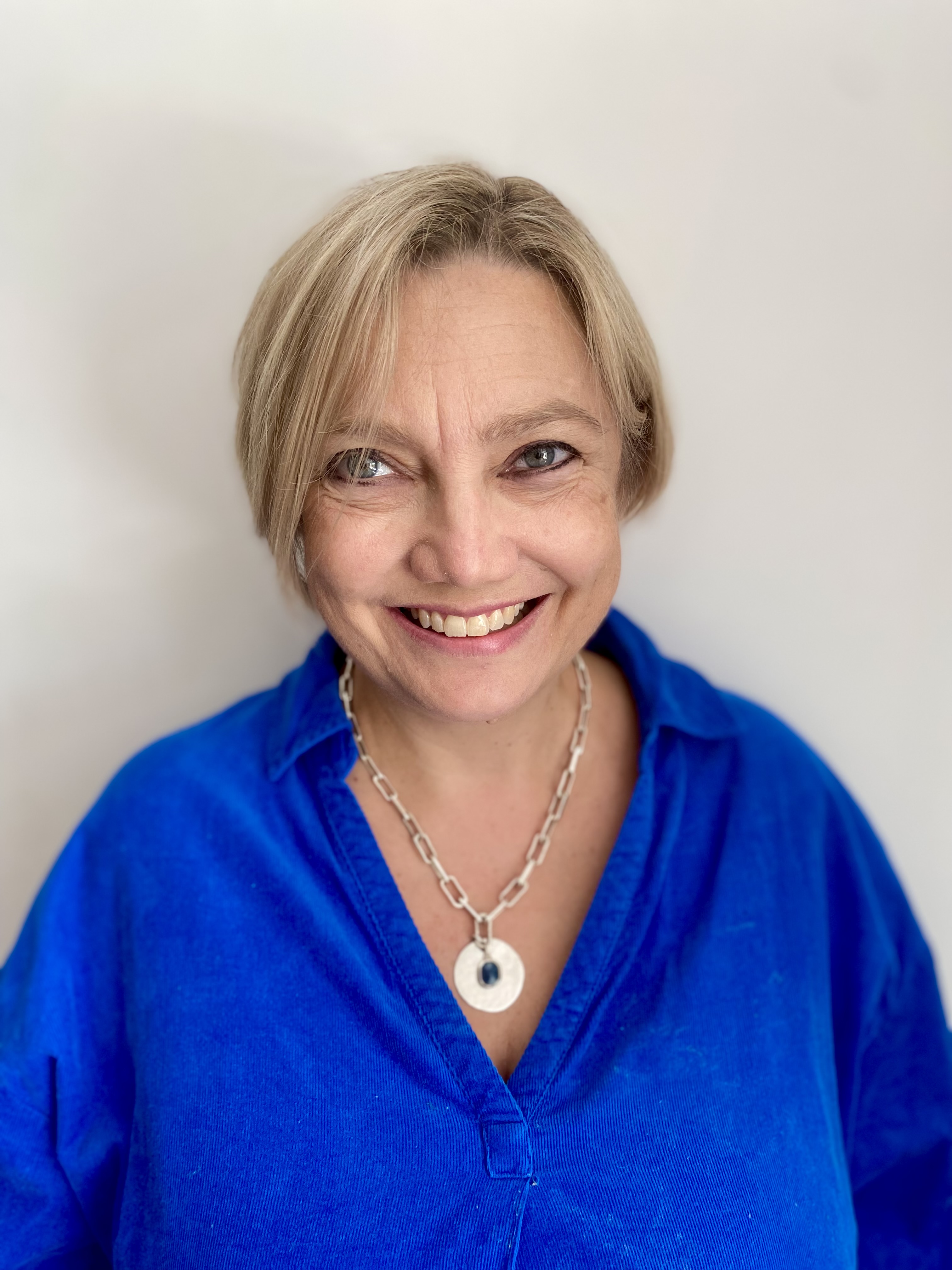 Head and shoulders photo of a woman with blond hair and wearing a blue shirt and gold chain necklace looking into the camera