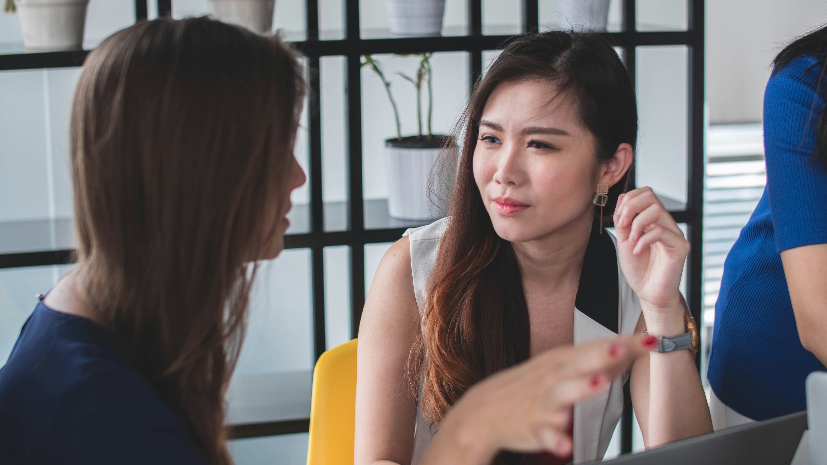 Two women sitting at a desk having a discussion with a laptop in front of them, in a modern office setting with shelves and plants in the background.