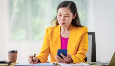 Person at office desk, looking at phone and writing on clipboard.