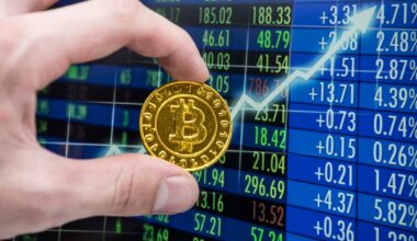 Hand holding coin with Bitcoin logo against a screen with a rising stock chart and tables with stock price data.