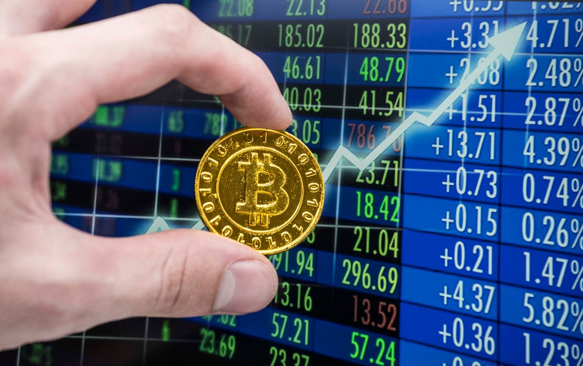 Hand holding coin with Bitcoin logo against a screen with a rising stock chart and tables with stock price data.