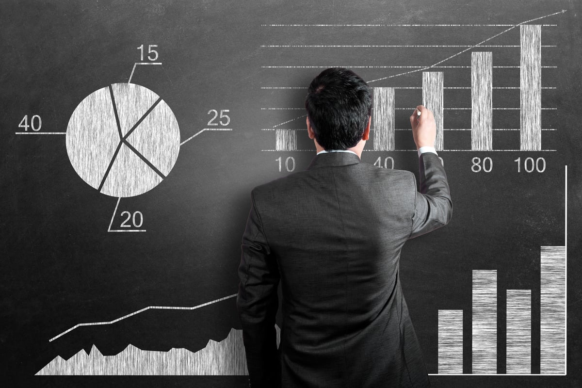 A person in a black suit draws stock-price charts on a blackboard.