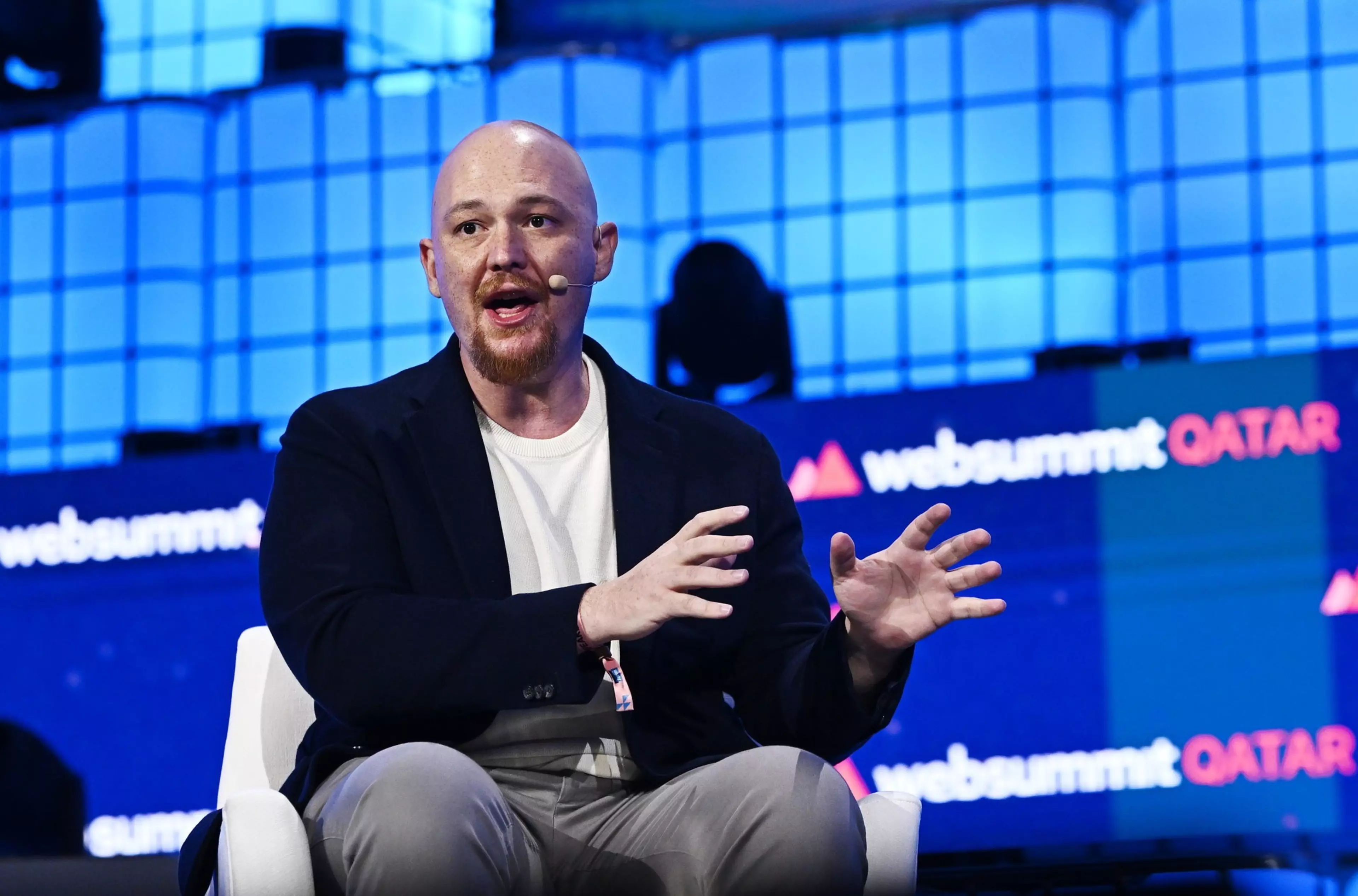 A bald man with a beard and headset mic is seated, gesturing with his hands while speaking at Web Summit Qatar, with the event name displayed on screens behind him.