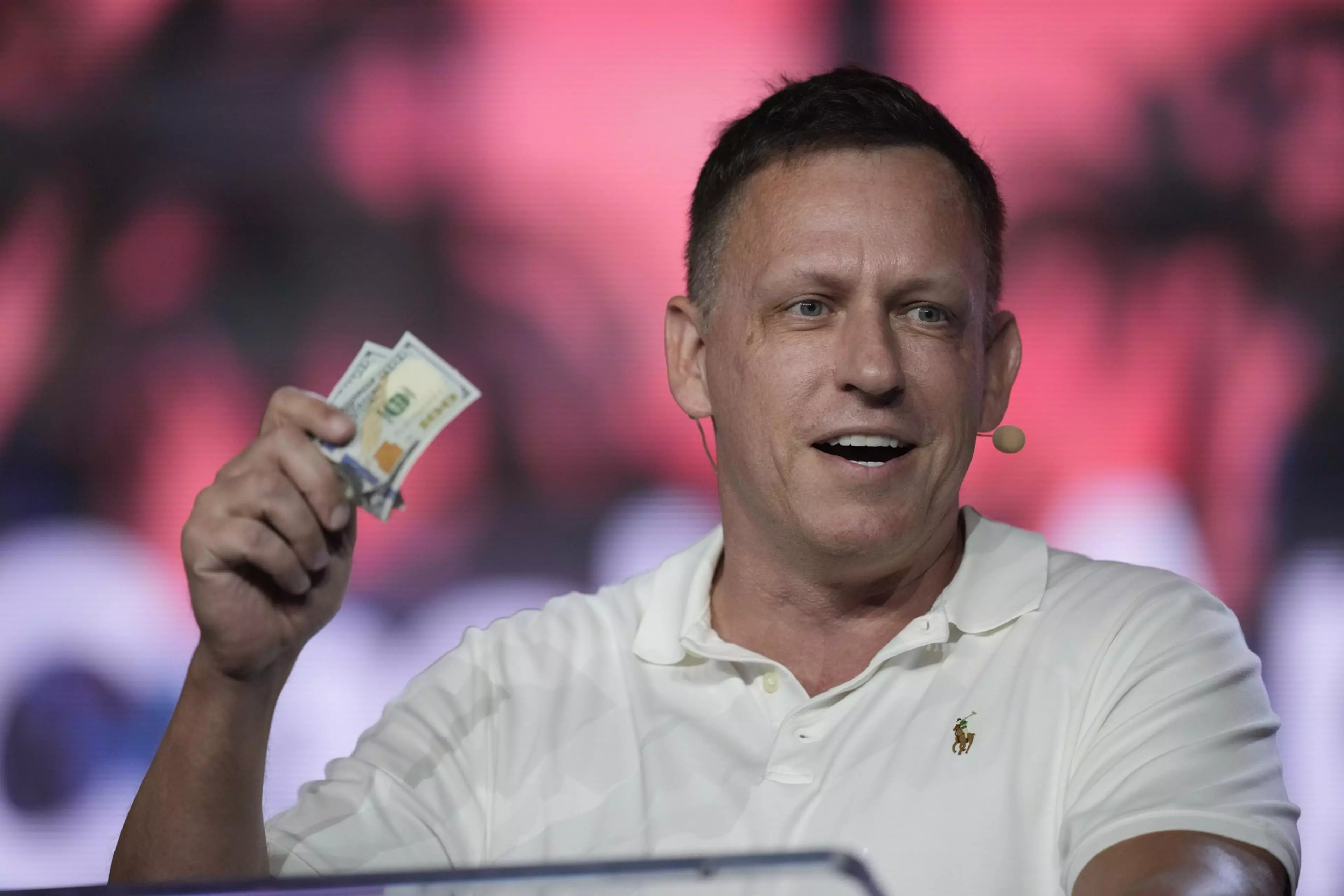 A man wearing a white polo shirt and a headset microphone holds up two $100 bills with a smile against a blurred red and black background.