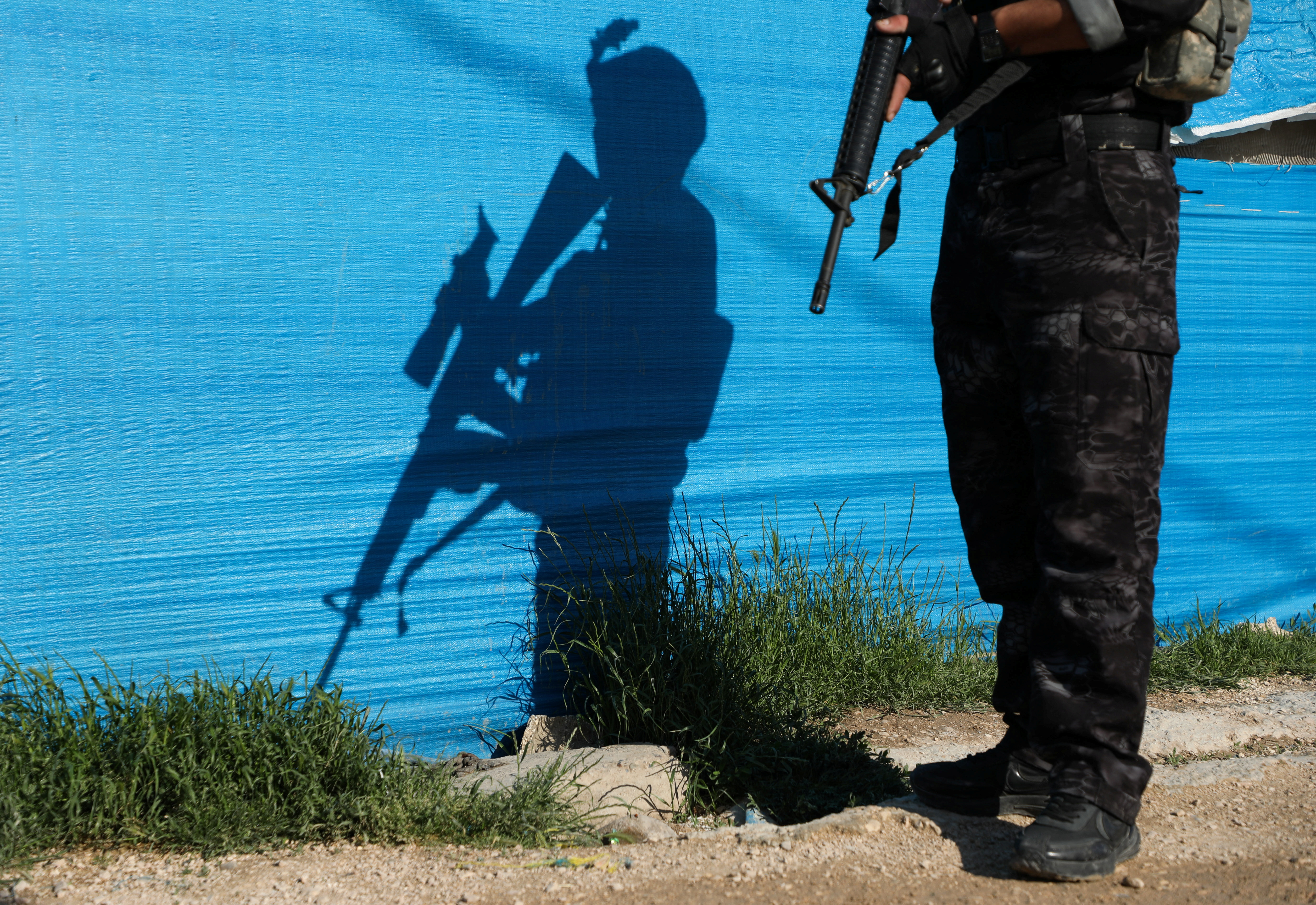 A person in camouflage pants holds a weapon, casting a shadow of themselves and their weapon on a bright blue tarp, during a security operation in Al-Roj camp, Syria.
