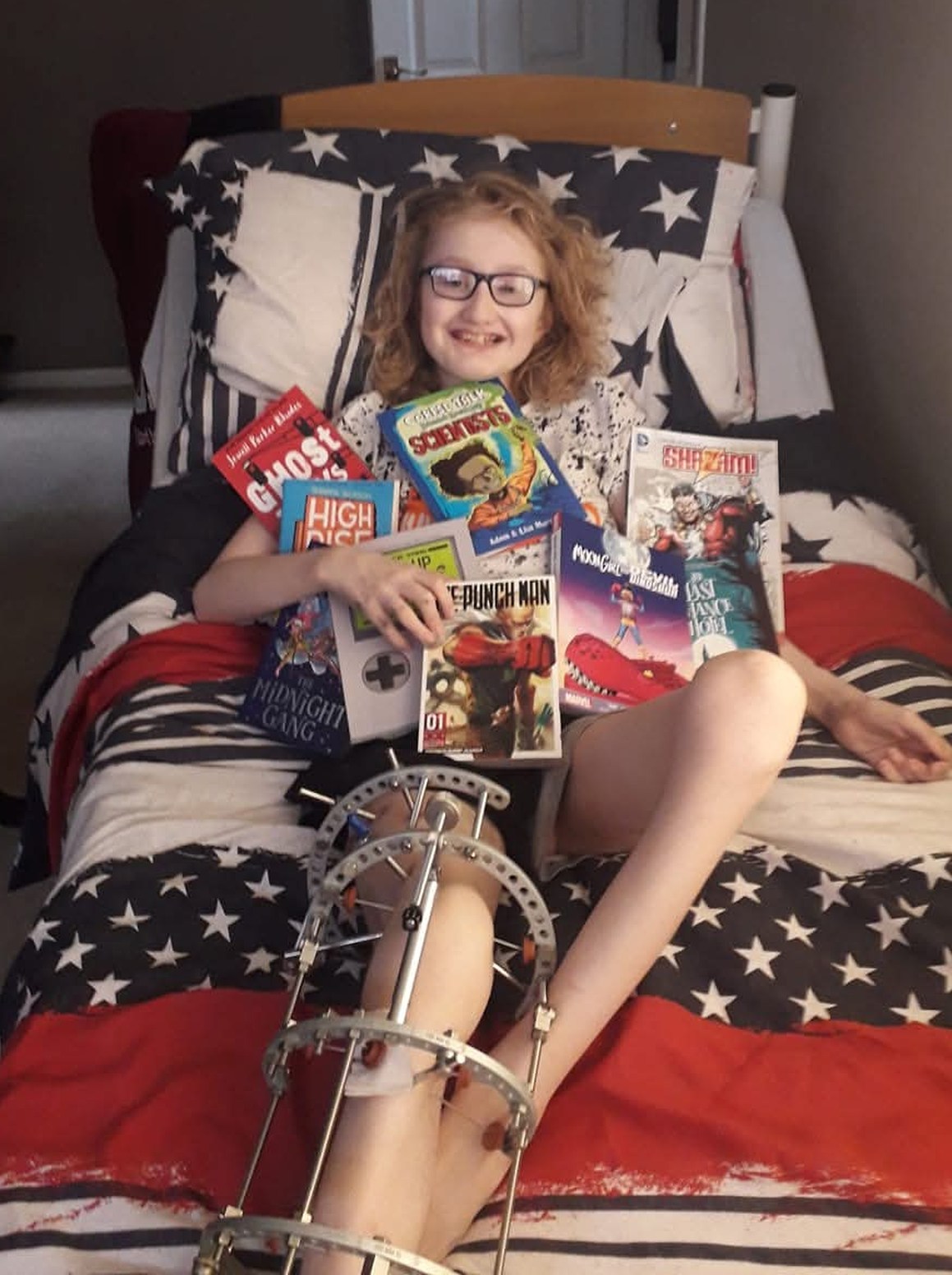 A young person with glasses and a leg in a frame, lying in bed, holding several books and smiling.
