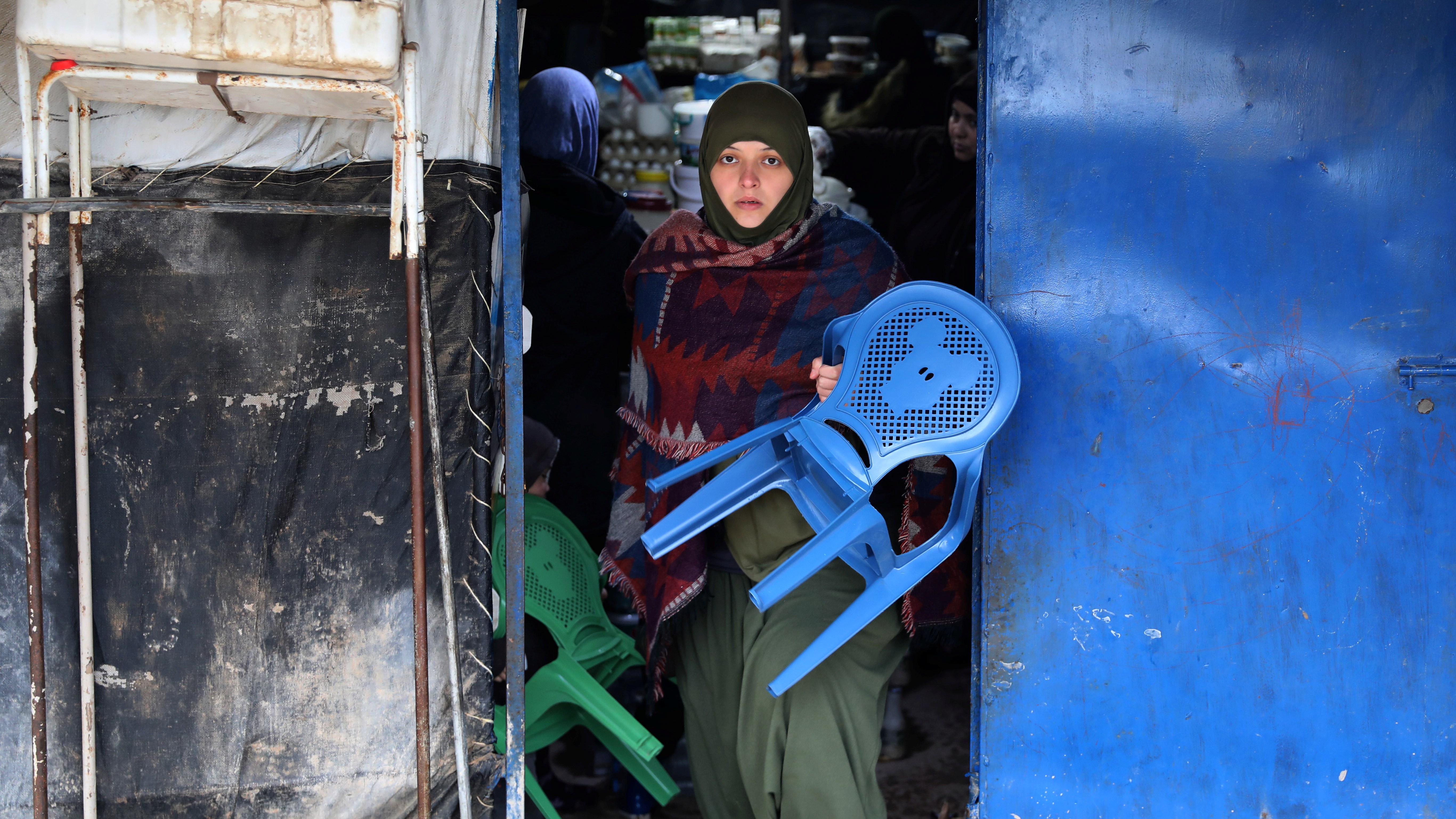 A woman wearing a hijab carries blue chairs out of a shop in al-Roj camp, Syria.