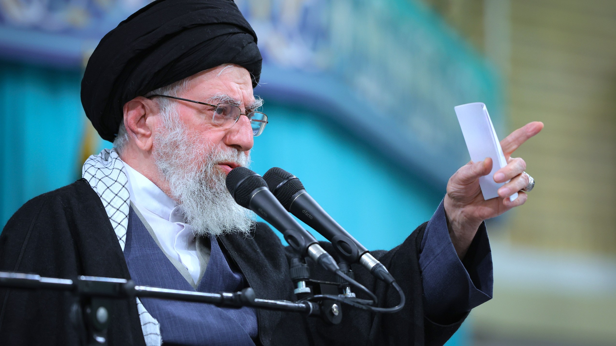 Supreme Leader Ayatollah Ali Khamenei speaks in a meeting, holding a small paper and gesturing with his right hand, with two microphones in front of him.