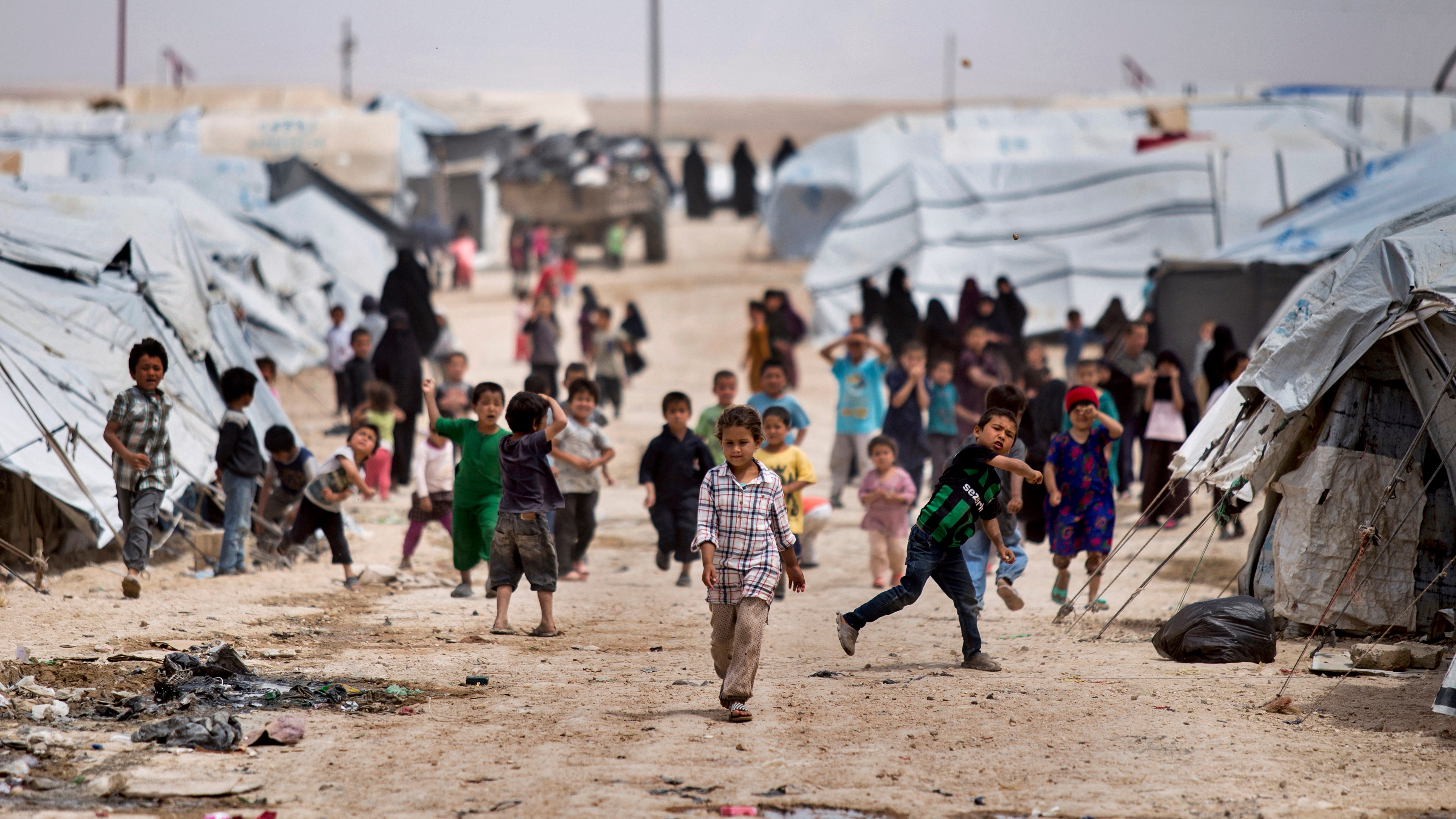 Children playing in a refugee camp with tents and debris in the background.