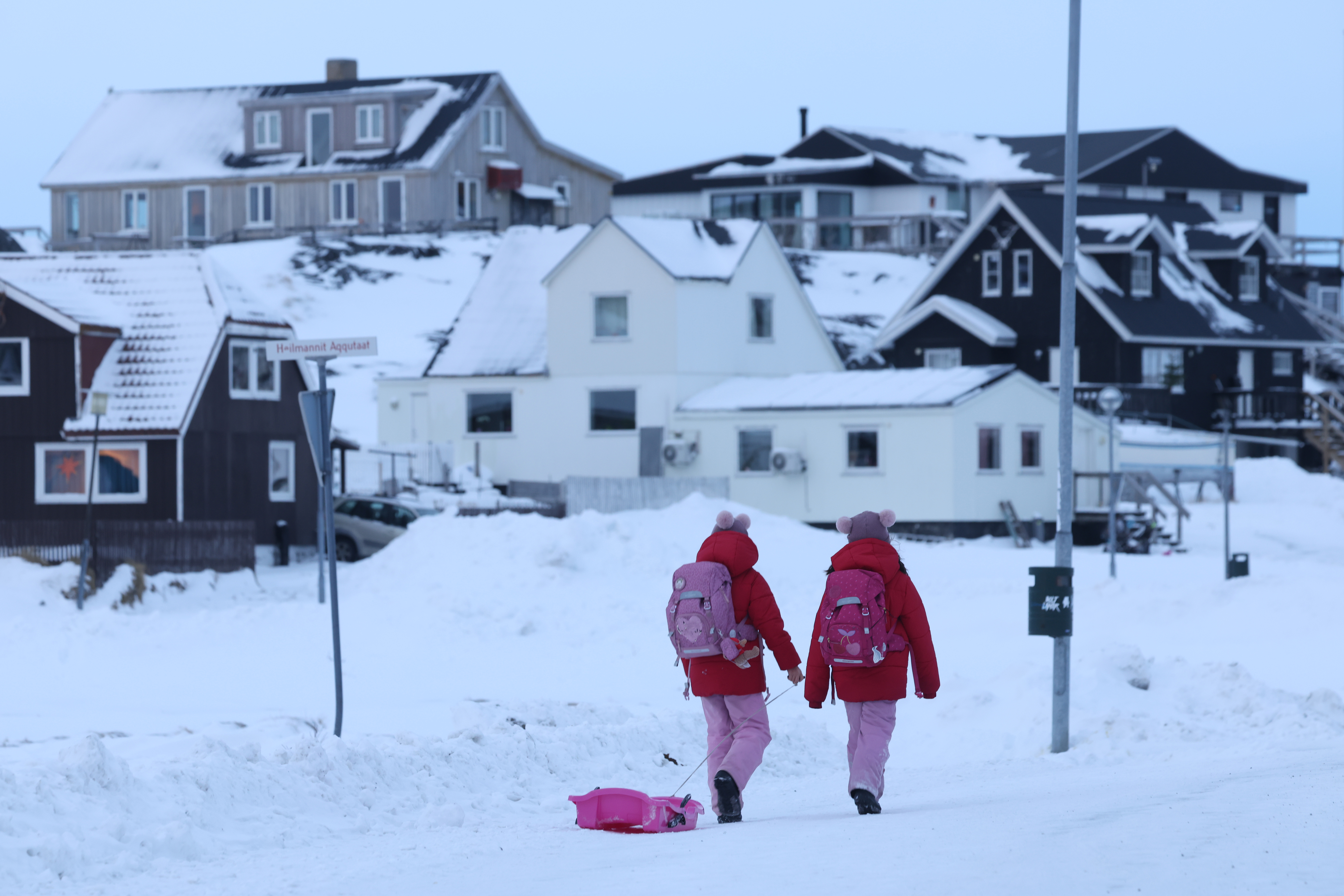 Two children in pink winter gear pull a sled down a snowy street in Nuuk, Greenland.