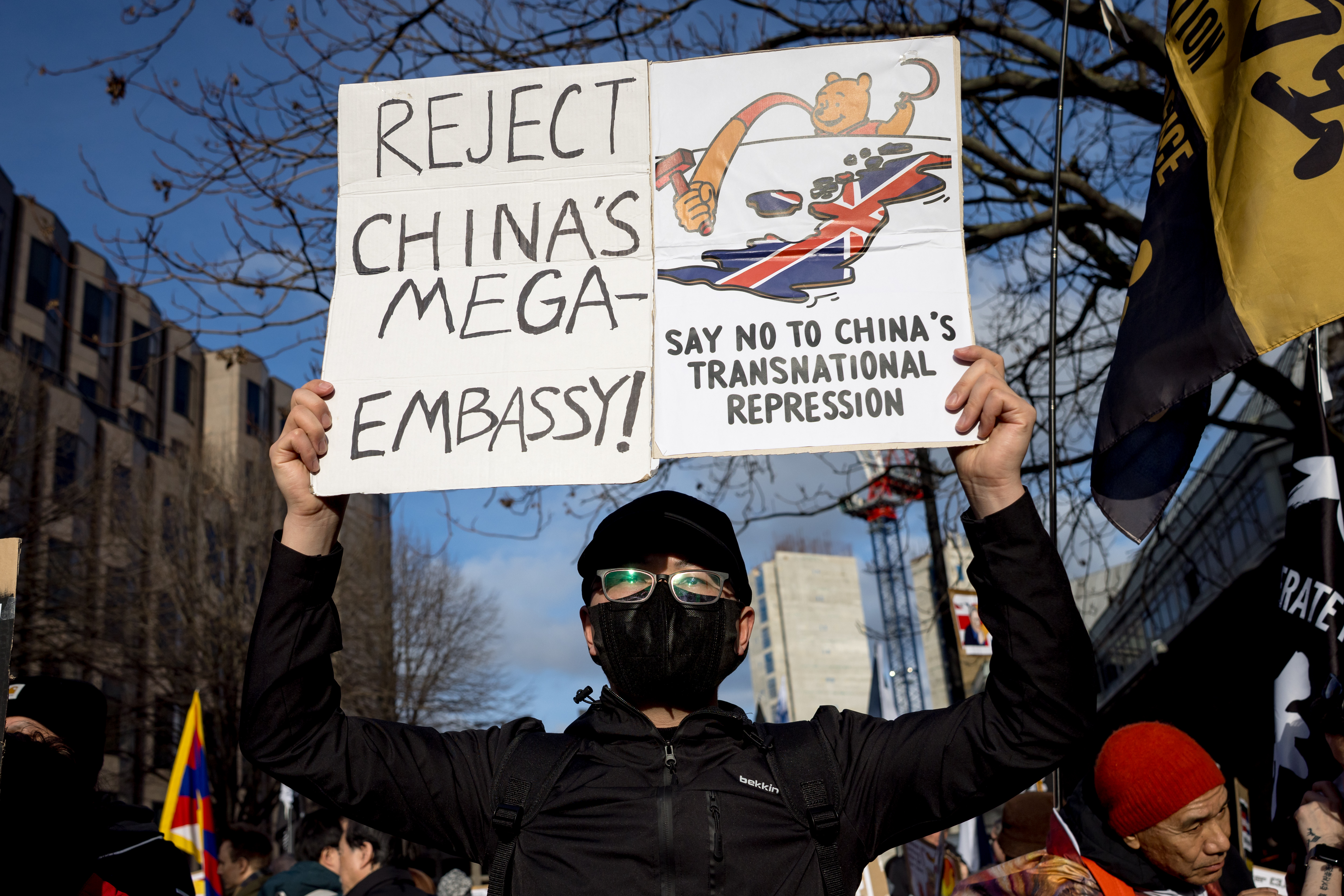 A person holding a protest sign that reads "REJECT CHINA'S MEGA-EMBASSY!" and "SAY NO TO CHINA'S TRANSNATIONAL REPRESSION."