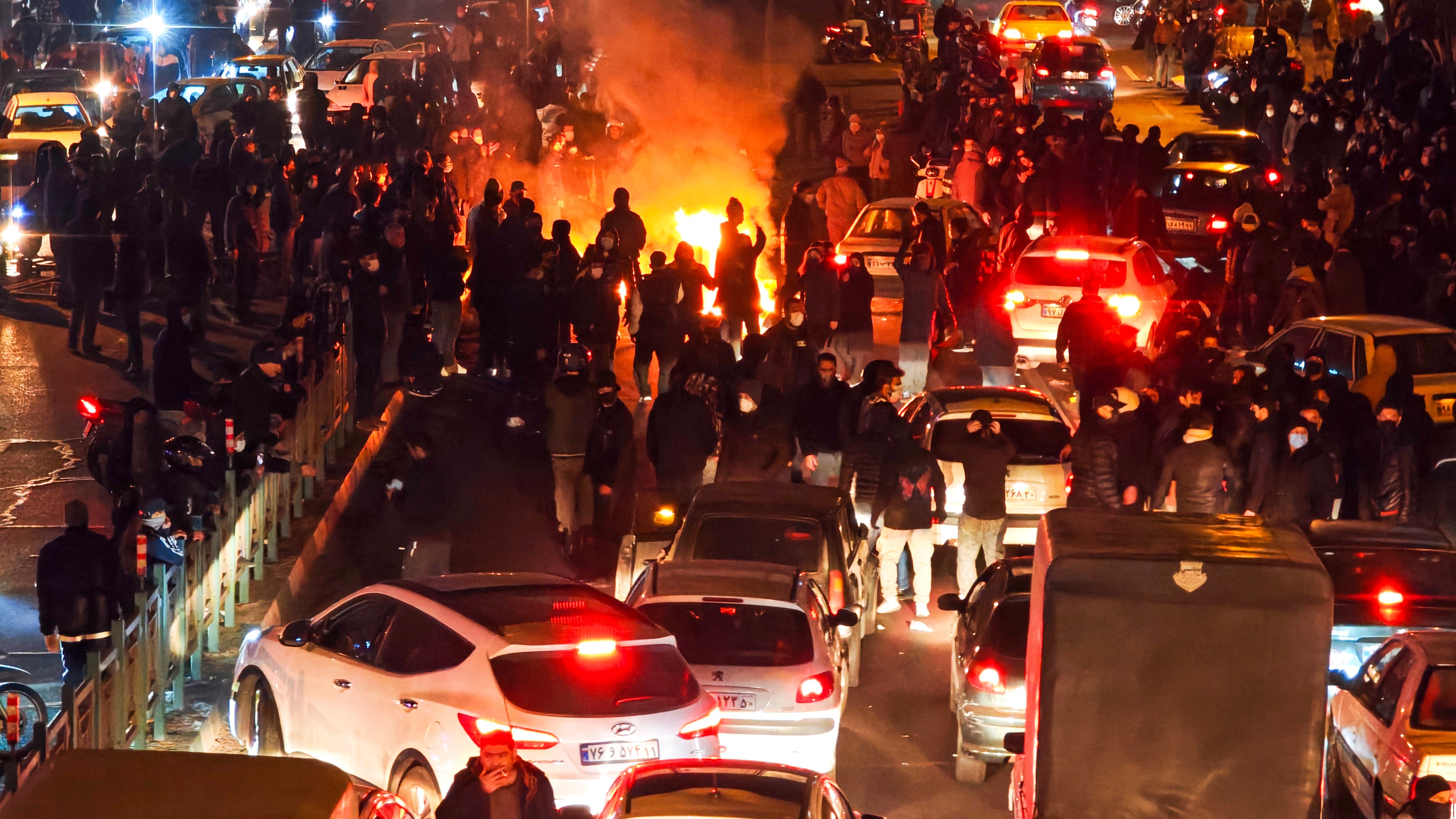 People protesting amid traffic and a large fire at night in Tehran, Iran.