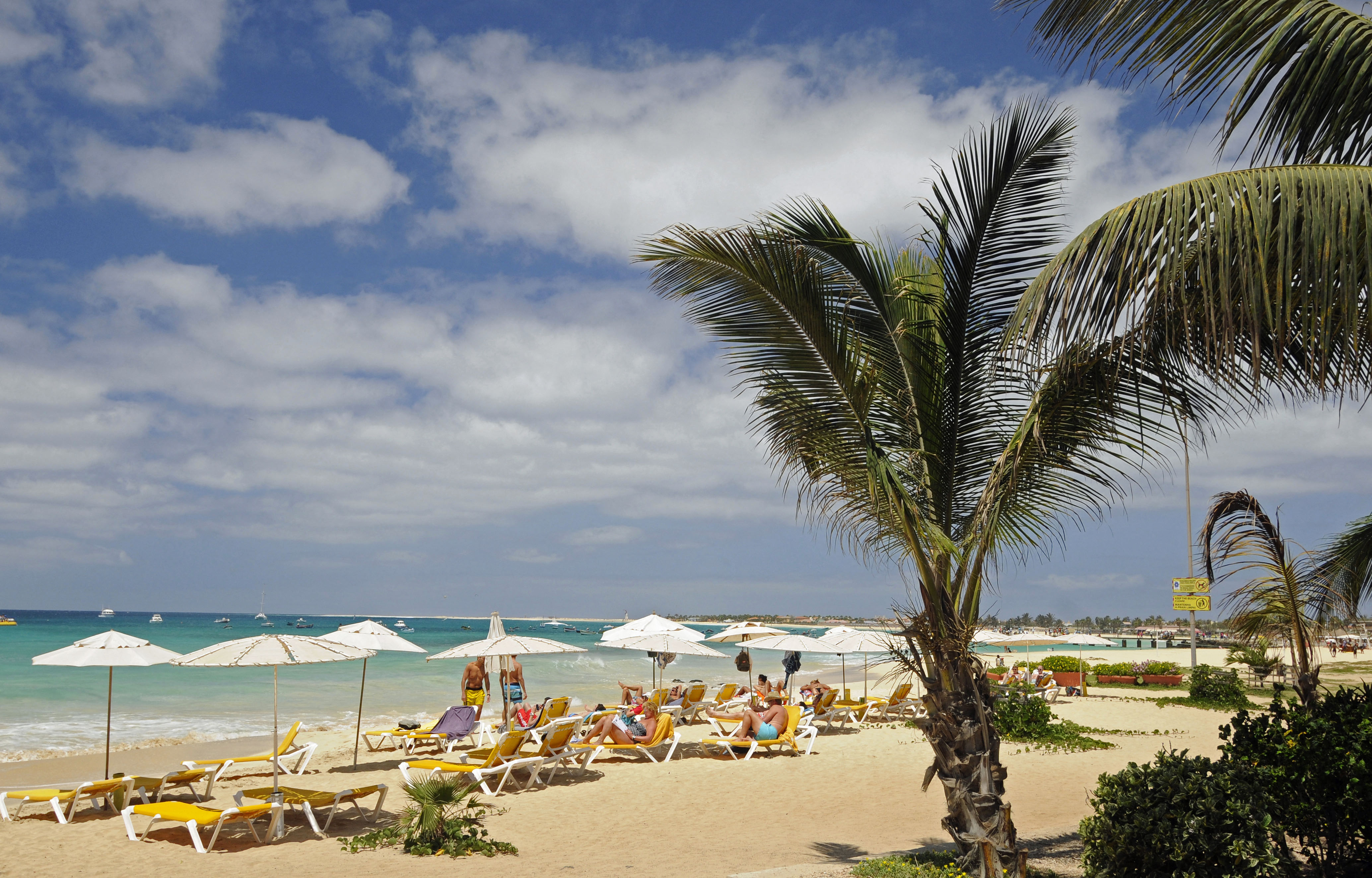 Santa Maria Beach on the island of Sal in Cape Verde, with people relaxing on lounge chairs under umbrellas, boats in the water, and palm trees.
