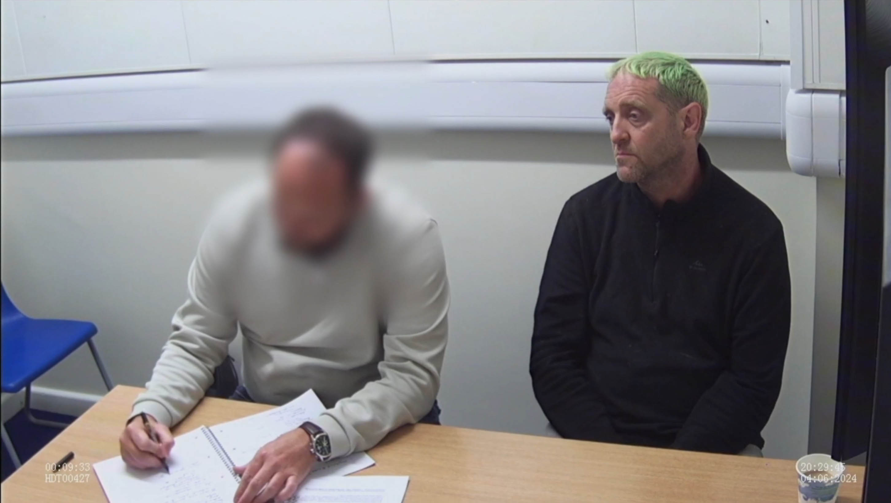 Robert Rhodes, with green hair, sits opposite an investigator who is writing in a notebook.