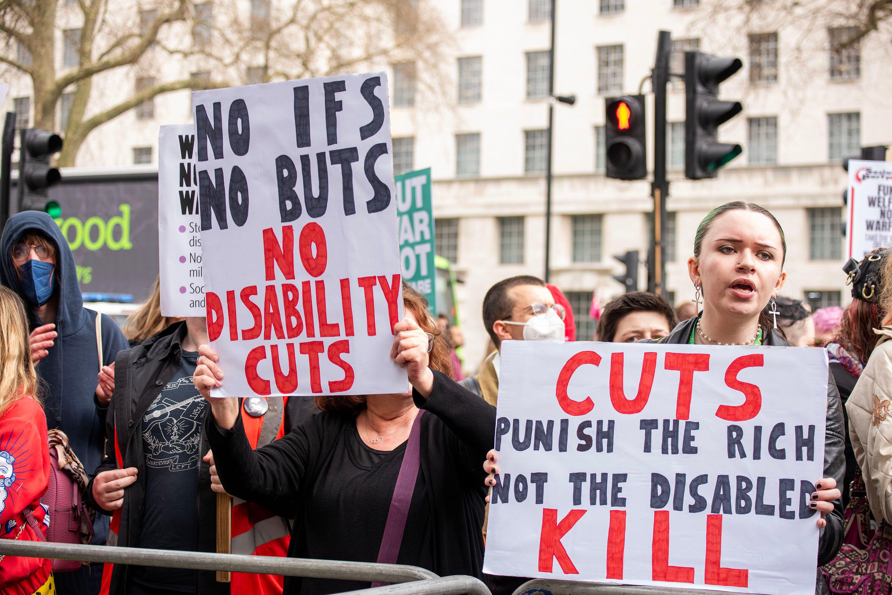 Protesters in London hold signs that read "No ifs, no buts, no disability cuts" and "Cuts punish the rich, not the disabled."
