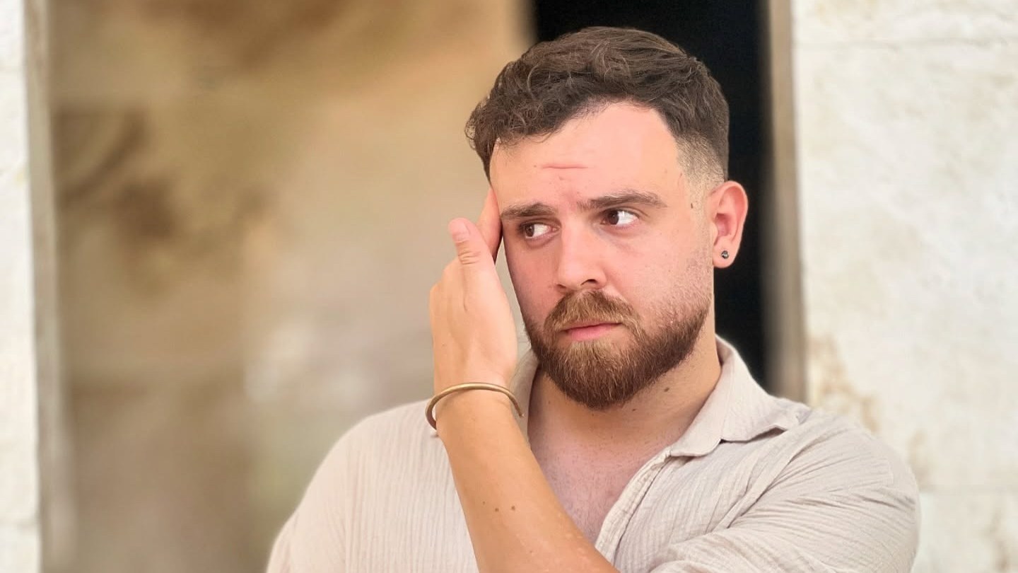 Erfan Soltani, a man with a beard, wearing a light-colored, long-sleeved shirt and a watch, looks to the right with his hand touching his temple.