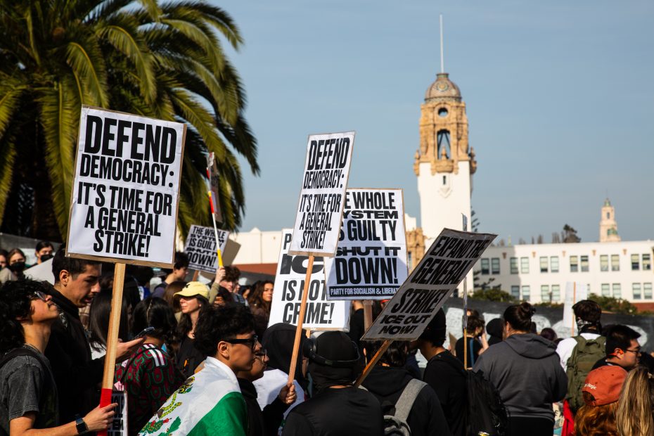 A group of protesters hold signs calling for a general strike and defending democracy, gathered outdoors near a tall clock tower and palm trees.