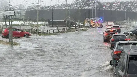 Fubar News Traffic queues along a flooded road as cars drive through standing water, with emergency vehicles flashing blue lights in the distance during heavy rain.