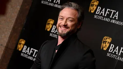 Getty Images McAvoy wearing a black coat standing in front of a BAFTA Scotland Awards backdrop with gold BAFTA logos and text on a black background